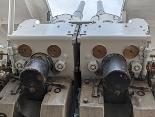 A close-up view of a dual-barrel artillery or anti-aircraft gun mounted on a ship or military vehicle. The metal components are painted in light gray with some areas exhibiting rust and wear. The barrels are parallel and centered in the image, with mechanical parts such as bolts and pistons visible around the base.