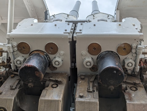 A close-up view of a dual-barrel artillery or anti-aircraft gun mounted on a ship or military vehicle. The metal components are painted in light gray with some areas exhibiting rust and wear. The barrels are parallel and centered in the image, with mechanical parts such as bolts and pistons visible around the base.