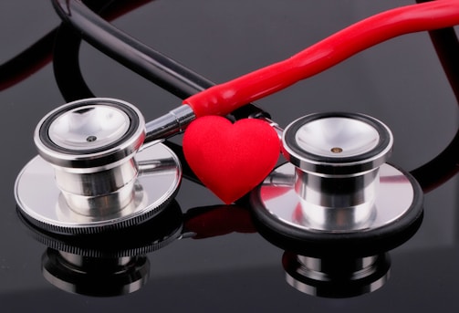 Two stethoscopes with shiny metallic diaphragms are arranged on a reflective black surface, with a small red heart placed in the center between them.
