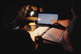 Two people studying or working together at a desk under a small desk lamp. One person is holding a pen, possibly taking notes in an open book, while the other is pointing at a laptop screen displaying text. The surrounding environment is dimly lit, with a focus on the workspace.