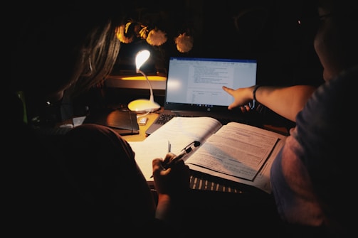 Two people studying or working together at a desk under a small desk lamp. One person is holding a pen, possibly taking notes in an open book, while the other is pointing at a laptop screen displaying text. The surrounding environment is dimly lit, with a focus on the workspace.