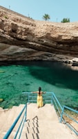 A person in a yellow dress is standing on a staircase that leads down into a natural water sinkhole surrounded by rocky walls. The water is a vibrant turquoise, and there is a palm tree visible above the rock wall in the background.