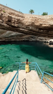 A person in a yellow dress is standing on a staircase that leads down into a natural water sinkhole surrounded by rocky walls. The water is a vibrant turquoise, and there is a palm tree visible above the rock wall in the background.