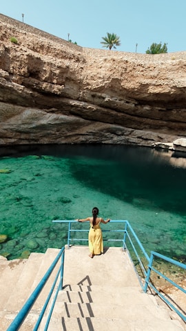 A person in a yellow dress is standing on a staircase that leads down into a natural water sinkhole surrounded by rocky walls. The water is a vibrant turquoise, and there is a palm tree visible above the rock wall in the background.