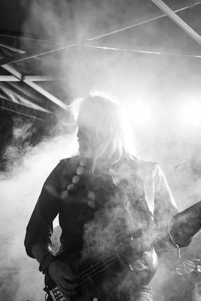A person is playing an electric guitar under bright stage lights, surrounded by smoke, creating a dramatic visual effect. The setting appears to be a live music performance with structural elements of the stage visible in the background.