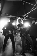 Two guitarists perform on stage under dramatic lighting, surrounded by smoke. The atmosphere is lively and energetic, with the musicians deeply engaged in their performance. The backdrop includes metal scaffolding, enhancing the concert setting.