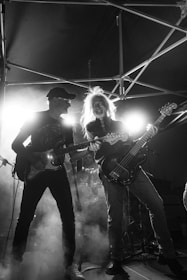 Two guitarists perform on stage under dramatic lighting, surrounded by smoke. The atmosphere is lively and energetic, with the musicians deeply engaged in their performance. The backdrop includes metal scaffolding, enhancing the concert setting.