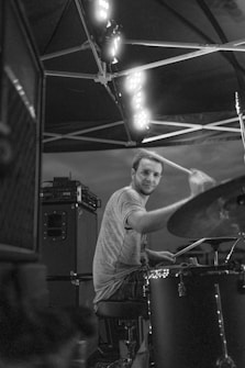 A black and white image featuring a person playing a drum set, with lights overhead illuminating the scene. The person appears focused and engaged in their music performance. The background shows part of a stage setup with speakers.