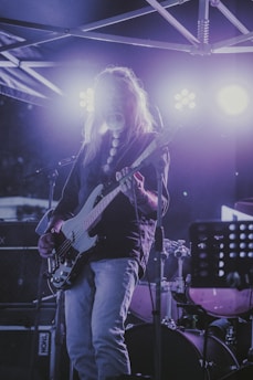 A musician with long hair is playing an electric guitar on stage under bright lights. There's a drum set in the background and the atmosphere suggests a concert or live performance. The lighting is predominantly purple with highlights around the musician.