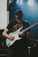 A guitarist passionately playing an electric guitar on stage under bright lights. The musician is wearing a cap and glasses, and appears to be deeply engaged in his performance.