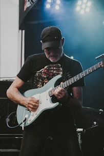 A guitarist passionately playing an electric guitar on stage under bright lights. The musician is wearing a cap and glasses, and appears to be deeply engaged in his performance.