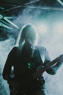 A musician with long hair plays an electric guitar on stage, illuminated by bright stage lights in a foggy atmosphere. The scene is dramatic with the lighting creating a high contrast effect, casting a silhouette of the musician.