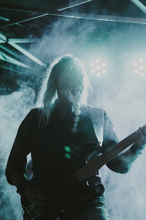 A musician with long hair plays an electric guitar on stage, illuminated by bright stage lights in a foggy atmosphere. The scene is dramatic with the lighting creating a high contrast effect, casting a silhouette of the musician.