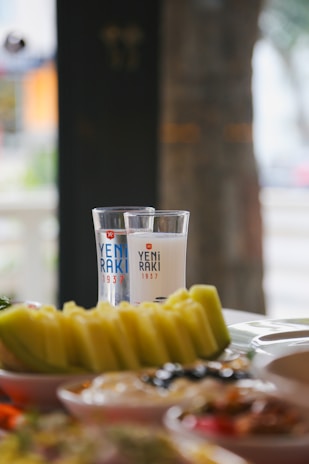 Two glasses of Yeni Raki with a selection of colorful appetizers and sliced fruits, likely melons, arranged on a table. The setting suggests a casual dining experience and has a blurred outdoor background.