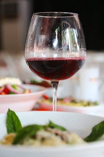 A glass of red wine is prominently positioned with a soft focus background of dishes featuring vibrant salads and greens. The glass reflects some light, adding a touch of elegance, and the table setting suggests a fine dining experience.