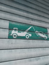 A green and white sign indicating a tow truck pulling a car, mounted on a corrugated metal shutter with a textured, weathered appearance.