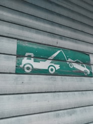 A green and white sign indicating a tow truck pulling a car, mounted on a corrugated metal shutter with a textured, weathered appearance.
