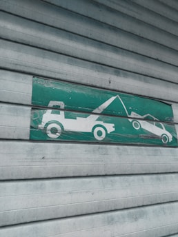 A green and white sign indicating a tow truck pulling a car, mounted on a corrugated metal shutter with a textured, weathered appearance.