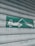 A green and white sign indicating a tow truck pulling a car, mounted on a corrugated metal shutter with a textured, weathered appearance.