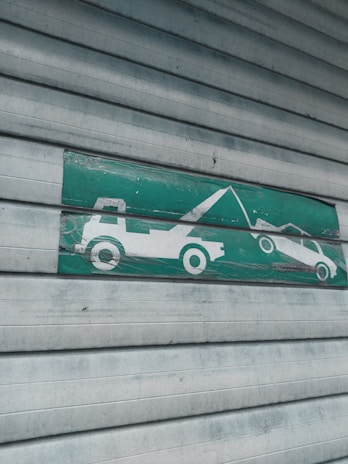 A green and white sign indicating a tow truck pulling a car, mounted on a corrugated metal shutter with a textured, weathered appearance.