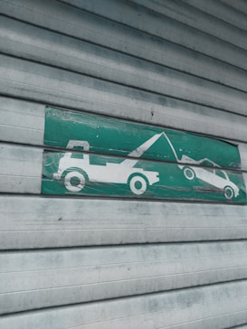 A green and white sign indicating a tow truck pulling a car, mounted on a corrugated metal shutter with a textured, weathered appearance.