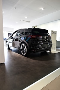 A sleek black Volkswagen ID.3 electric car is displayed inside a modern showroom with bright lighting. The vehicle is positioned on a raised platform beneath a white ceiling, with reflective surfaces highlighting its glossy finish. Nearby, a charging station marked with the Ohme logo is visible.