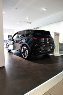 A sleek black Volkswagen ID.3 electric car is displayed inside a modern showroom with bright lighting. The vehicle is positioned on a raised platform beneath a white ceiling, with reflective surfaces highlighting its glossy finish. Nearby, a charging station marked with the Ohme logo is visible.