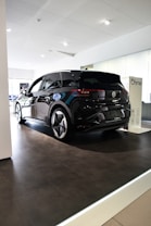 A sleek black Volkswagen ID.3 electric car is displayed inside a modern showroom with bright lighting. The vehicle is positioned on a raised platform beneath a white ceiling, with reflective surfaces highlighting its glossy finish. Nearby, a charging station marked with the Ohme logo is visible.