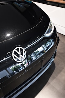 A close-up of the front section of a black Volkswagen car, prominently displaying the Volkswagen logo on the hood. The car's sleek design and reflective surface are evident. The environment is bright, suggesting indoor lighting, with the vehicle positioned on a showroom floor.