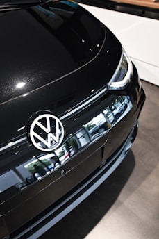 A close-up of the front section of a black Volkswagen car, prominently displaying the Volkswagen logo on the hood. The car's sleek design and reflective surface are evident. The environment is bright, suggesting indoor lighting, with the vehicle positioned on a showroom floor.