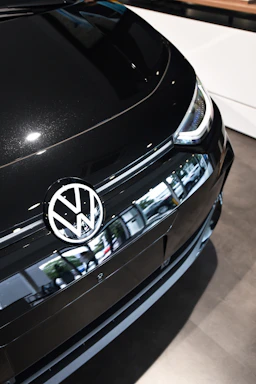 A close-up of the front section of a black Volkswagen car, prominently displaying the Volkswagen logo on the hood. The car's sleek design and reflective surface are evident. The environment is bright, suggesting indoor lighting, with the vehicle positioned on a showroom floor.