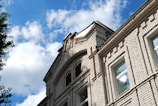 A close-up of a brick building facade with detailed masonry work, featuring square windows and an arched design near the roofline. The sky is blue with scattered white clouds, and part of a tree is visible on the left.