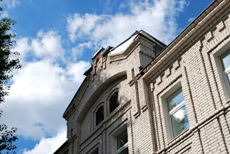 A close-up of a brick building facade with detailed masonry work, featuring square windows and an arched design near the roofline. The sky is blue with scattered white clouds, and part of a tree is visible on the left.