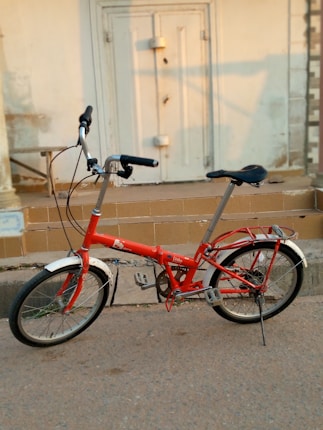 A red folding bicycle is parked on the side of a paved road near some tiled steps leading to a white door with a latch. The bicycle has a black seat, black handles, and silver handlebars. The front and back wheels both have fenders, and there is a small kickstand supporting the bicycle.