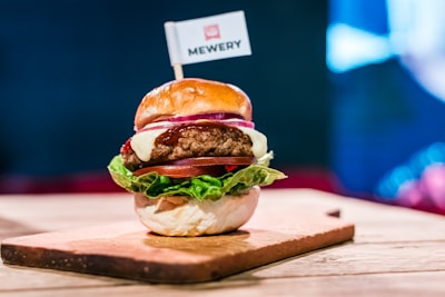 A gourmet hamburger is presented on a wooden board. The burger includes a soft, shiny bun, a juicy beef patty, melted cheese, slices of fresh tomato and red onion, and crisp green lettuce. A small flag with the word 'MEWERY' is placed on top of the bun. The background appears to be a blurred multi-colored light, providing a vibrant yet cozy setting.