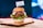A gourmet hamburger is presented on a wooden board. The burger includes a soft, shiny bun, a juicy beef patty, melted cheese, slices of fresh tomato and red onion, and crisp green lettuce. A small flag with the word 'MEWERY' is placed on top of the bun. The background appears to be a blurred multi-colored light, providing a vibrant yet cozy setting.