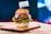 A gourmet hamburger is presented on a wooden board. The burger includes a soft, shiny bun, a juicy beef patty, melted cheese, slices of fresh tomato and red onion, and crisp green lettuce. A small flag with the word 'MEWERY' is placed on top of the bun. The background appears to be a blurred multi-colored light, providing a vibrant yet cozy setting.