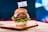 A gourmet hamburger is presented on a wooden board. The burger includes a soft, shiny bun, a juicy beef patty, melted cheese, slices of fresh tomato and red onion, and crisp green lettuce. A small flag with the word 'MEWERY' is placed on top of the bun. The background appears to be a blurred multi-colored light, providing a vibrant yet cozy setting.