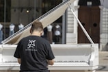 A person wearing a black t-shirt with a graphic design and website URL is playing a white grand piano. The piano is set outdoors with a backdrop of glass windows displaying mannequins dressed in various outfits. There is a large wooden door in the background.