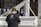 A person wearing a black t-shirt with a graphic design and website URL is playing a white grand piano. The piano is set outdoors with a backdrop of glass windows displaying mannequins dressed in various outfits. There is a large wooden door in the background.