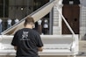 A person wearing a black t-shirt with a graphic design and website URL is playing a white grand piano. The piano is set outdoors with a backdrop of glass windows displaying mannequins dressed in various outfits. There is a large wooden door in the background.