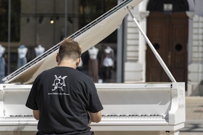 A person wearing a black t-shirt with a graphic design and website URL is playing a white grand piano. The piano is set outdoors with a backdrop of glass windows displaying mannequins dressed in various outfits. There is a large wooden door in the background.