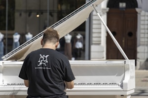 A person wearing a black t-shirt with a graphic design and website URL is playing a white grand piano. The piano is set outdoors with a backdrop of glass windows displaying mannequins dressed in various outfits. There is a large wooden door in the background.