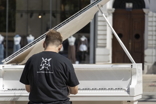 A person wearing a black t-shirt with a graphic design and website URL is playing a white grand piano. The piano is set outdoors with a backdrop of glass windows displaying mannequins dressed in various outfits. There is a large wooden door in the background.