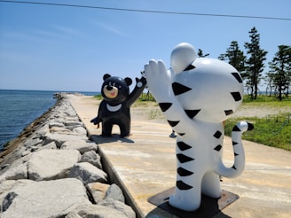 Two cartoon-like statues of animals located on a pathway by the sea, with a bear on the left and a tiger on the right. The bear is black with a cheerful expression and is raising one paw, while the white tiger features black stripes and is also waving. They are outdoors with a clear blue sky and trees in the background. The pathway runs next to a body of water and is lined with large rocks.