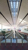 A spacious, well-lit airport terminal with high ceilings featuring parallel lines that create a modern and clean aesthetic. An empty moving walkway stretches forward, flanked by columns and glass barriers. A lone traveler walks down the walkway, surrounded by signs and pathways leading to different parts of the terminal.