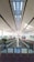 A spacious, well-lit airport terminal with high ceilings featuring parallel lines that create a modern and clean aesthetic. An empty moving walkway stretches forward, flanked by columns and glass barriers. A lone traveler walks down the walkway, surrounded by signs and pathways leading to different parts of the terminal.