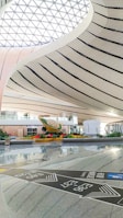 The spacious interior showcases a modern architectural design with sweeping, curved lines and a large, lattice-patterned skylight allowing natural light to flood the space. In the center, there is a striking sculpture surrounded by greenery, which adds a touch of nature to the sleek, contemporary setting. Signs with directional information are visible on the polished floor.