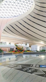 The spacious interior showcases a modern architectural design with sweeping, curved lines and a large, lattice-patterned skylight allowing natural light to flood the space. In the center, there is a striking sculpture surrounded by greenery, which adds a touch of nature to the sleek, contemporary setting. Signs with directional information are visible on the polished floor.