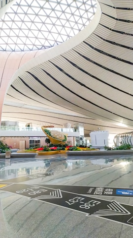 The spacious interior showcases a modern architectural design with sweeping, curved lines and a large, lattice-patterned skylight allowing natural light to flood the space. In the center, there is a striking sculpture surrounded by greenery, which adds a touch of nature to the sleek, contemporary setting. Signs with directional information are visible on the polished floor.